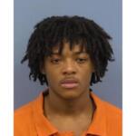 Mugshot of a young man with short dreadlocks wearing an orange jail uniform, facing the camera against a gray background.