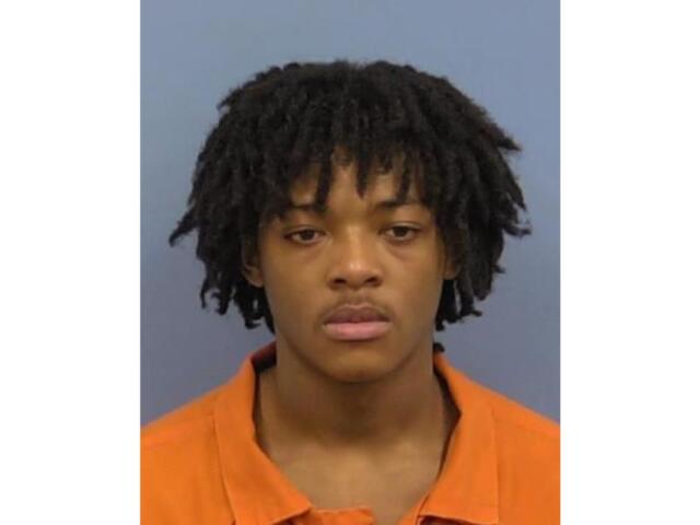 Mugshot of a young man with short dreadlocks wearing an orange jail uniform, facing the camera against a gray background.