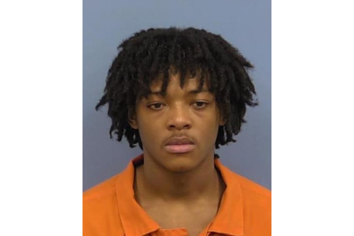 Mugshot of a young man with short dreadlocks wearing an orange jail uniform, facing the camera against a gray background.