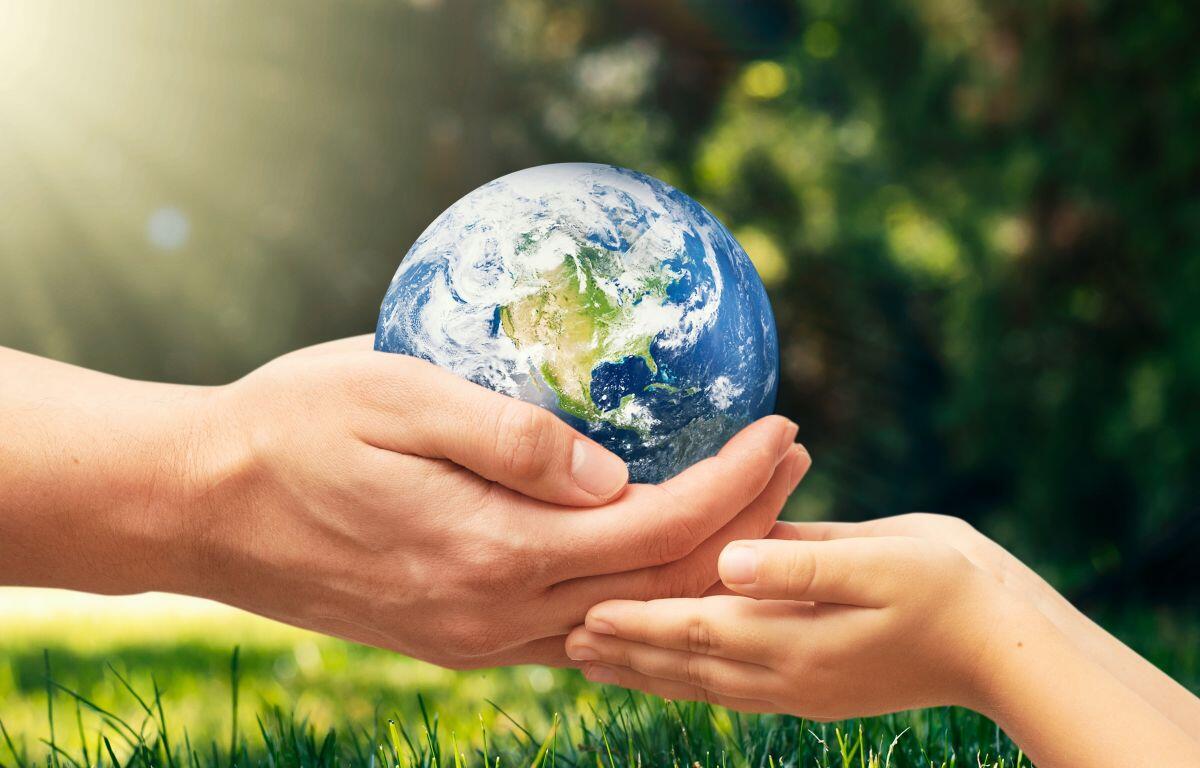 Two pairs of hands, adult and child, gently cradle a globe Earth outdoors in sunlight and grass.