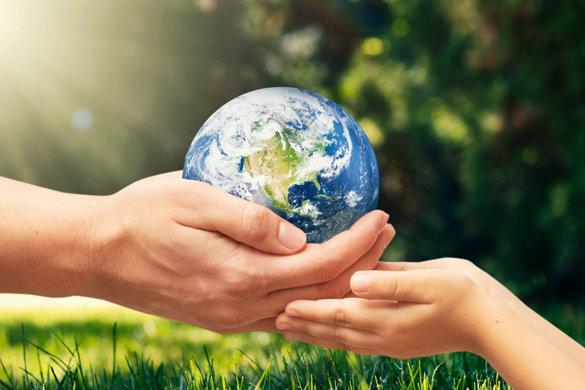 Two pairs of hands, adult and child, gently cradle a globe Earth outdoors in sunlight and grass.