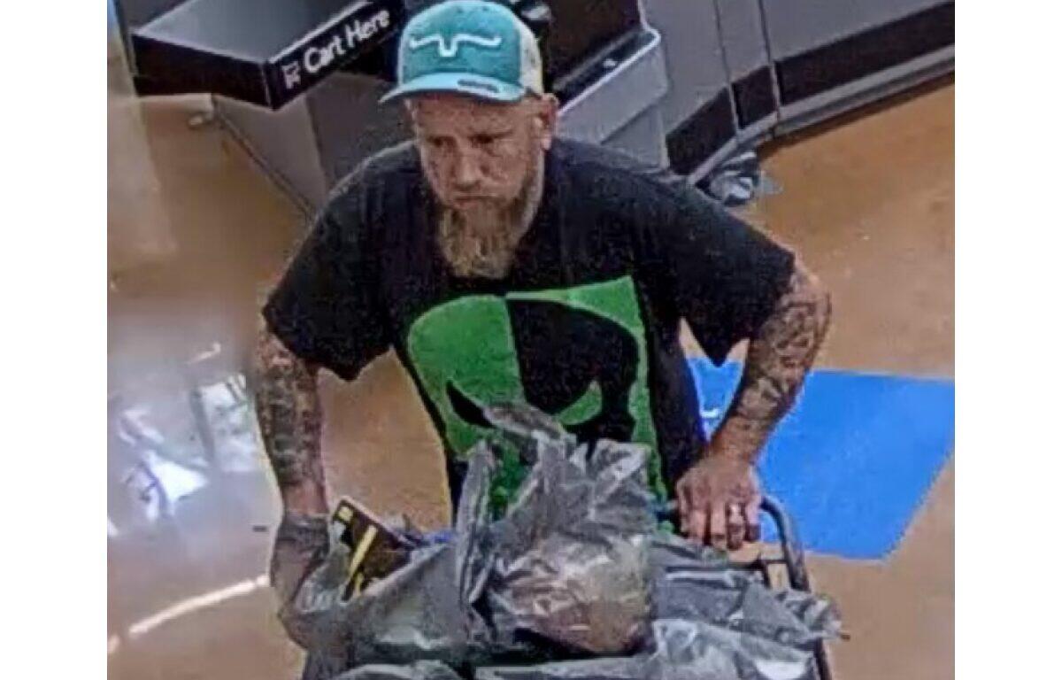 Bearded man wearing a teal cap and black graphic shirt pushes a shopping cart piled with plastic bags in a store aisle.
