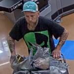 Bearded man wearing a teal cap and black graphic shirt pushes a shopping cart piled with plastic bags in a store aisle.