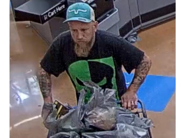 Bearded man wearing a teal cap and black graphic shirt pushes a shopping cart piled with plastic bags in a store aisle.