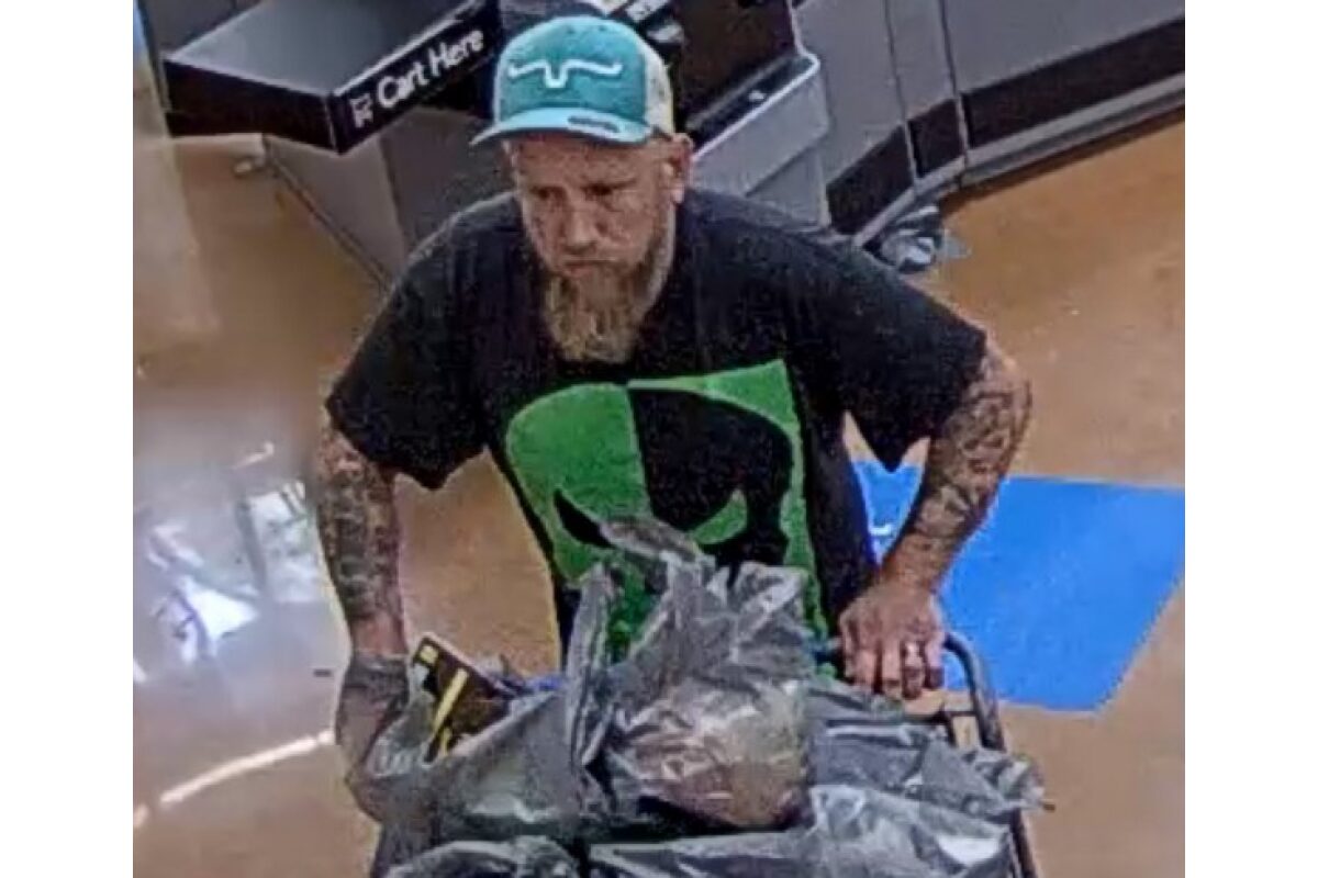Bearded man wearing a teal cap and black graphic shirt pushes a shopping cart piled with plastic bags in a store aisle.