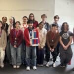 Group of diverse students in a hallway, wearing medals, posing with a tall trophy.