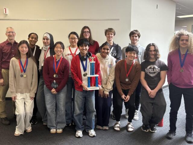 Group of diverse students in a hallway, wearing medals, posing with a tall trophy.