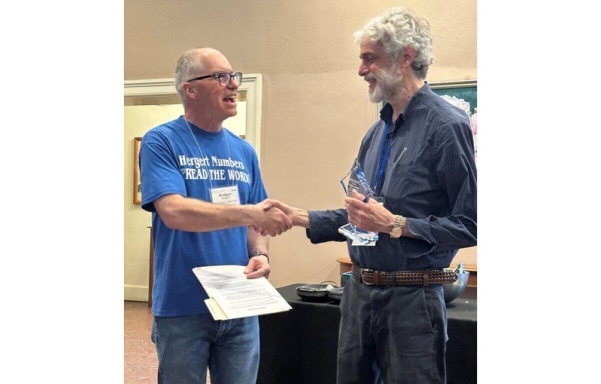 Two men shake hands during an award ceremony; the left man wears a blue T‑shirt and holds a certificate, the right man in a dark shirt holds a glass trophy.