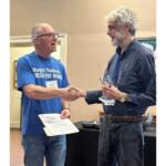 Two men shake hands during an award ceremony; the left man wears a blue T‑shirt and holds a certificate, the right man in a dark shirt holds a glass trophy.
