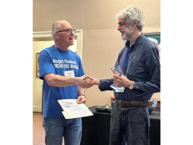 Two men shake hands during an award ceremony; the left man wears a blue T‑shirt and holds a certificate, the right man in a dark shirt holds a glass trophy.