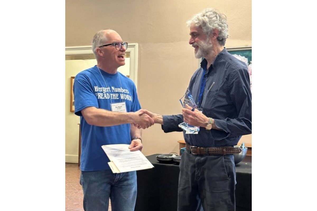 Two men shake hands during an award ceremony; the left man wears a blue T‑shirt and holds a certificate, the right man in a dark shirt holds a glass trophy.