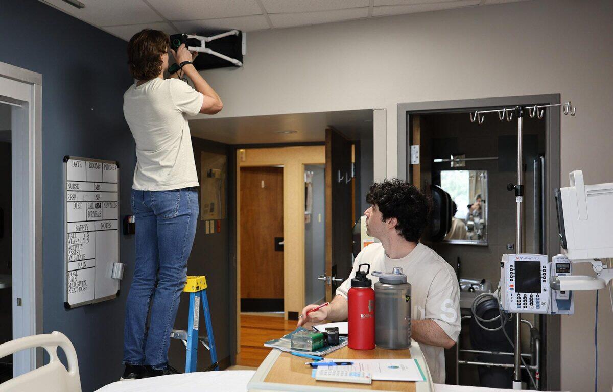 A person on a step ladder attaches equipment to the ceiling while another person sits at a bedside table marking notes in a hospital room.