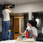 A person on a step ladder attaches equipment to the ceiling while another person sits at a bedside table marking notes in a hospital room.