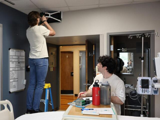 A person on a step ladder attaches equipment to the ceiling while another person sits at a bedside table marking notes in a hospital room.