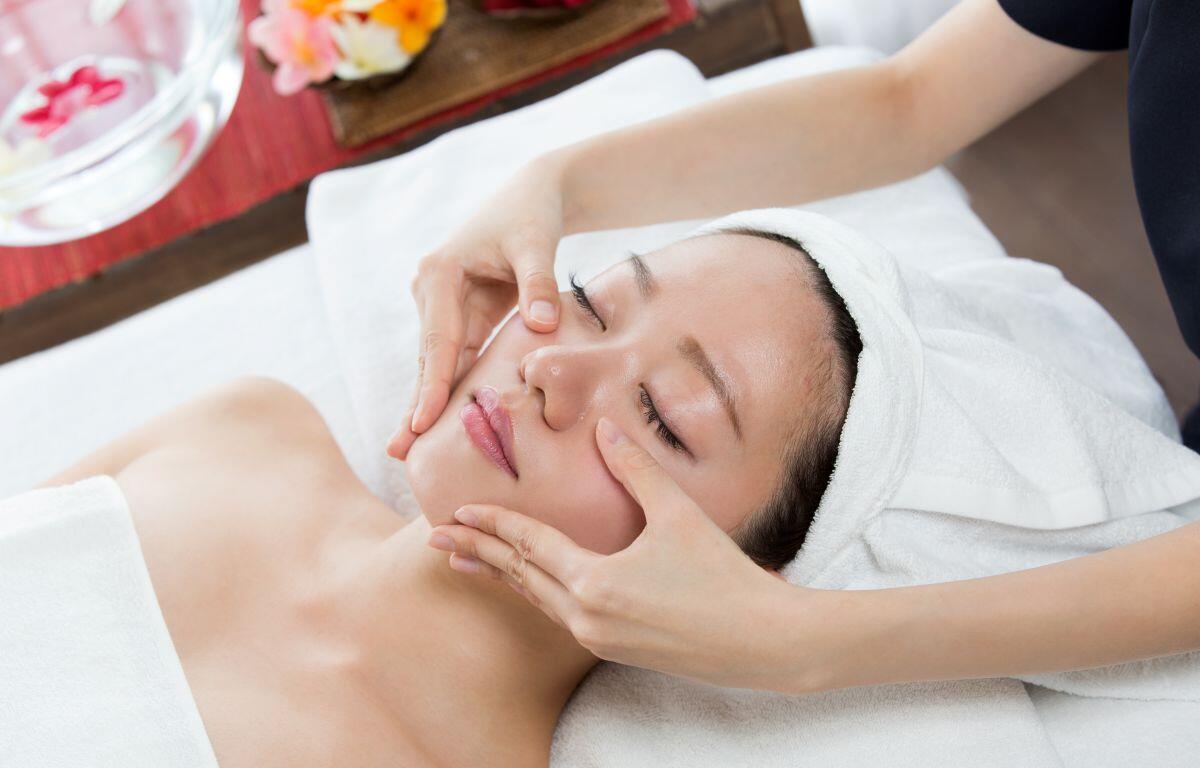 Spa facial treatment: woman lying with towel wrapped head while therapist gently massages cheeks and jaw.