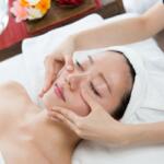 Spa facial treatment: woman lying with towel wrapped head while therapist gently massages cheeks and jaw.