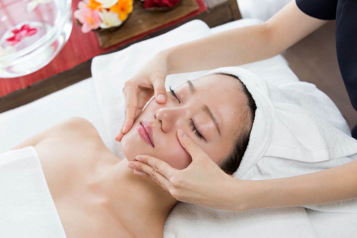 Spa facial treatment: woman lying with towel wrapped head while therapist gently massages cheeks and jaw.