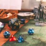 Tabletop RPG scene with a black twenty-sided die and several blue and red dice scattered on a gaming map, plus fantasy minis in the background.
