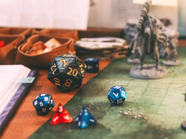 Tabletop RPG scene with a black twenty-sided die and several blue and red dice scattered on a gaming map, plus fantasy minis in the background.