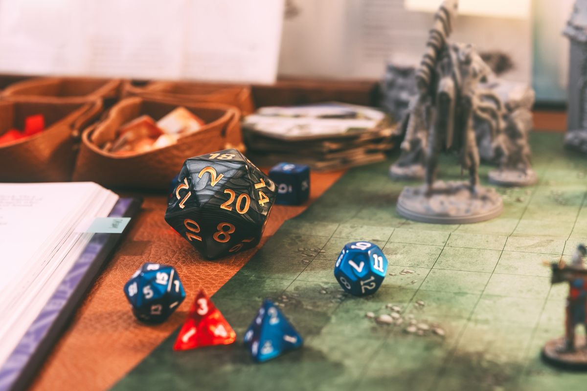 Tabletop RPG scene with a black twenty-sided die and several blue and red dice scattered on a gaming map, plus fantasy minis in the background.