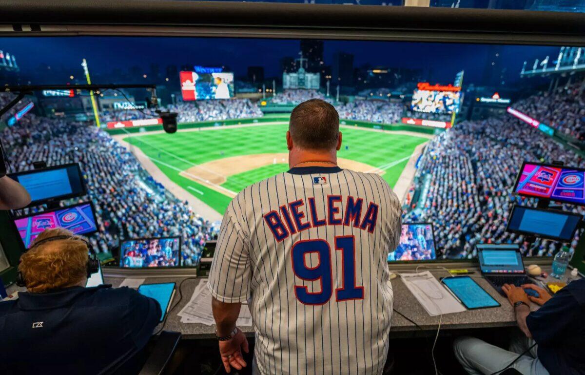 Man wearing a 'BIELEMA 91' pinstriped jersey in a broadcast booth, overlooking a packed baseball stadium at night.