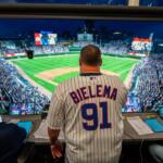 Man wearing a 'BIELEMA 91' pinstriped jersey in a broadcast booth, overlooking a packed baseball stadium at night.