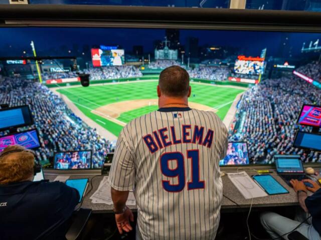 Man wearing a 'BIELEMA 91' pinstriped jersey in a broadcast booth, overlooking a packed baseball stadium at night.