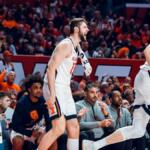 Illinois basketball players in white uniforms cheer on the sideline during a game, crowd in orange in the background.