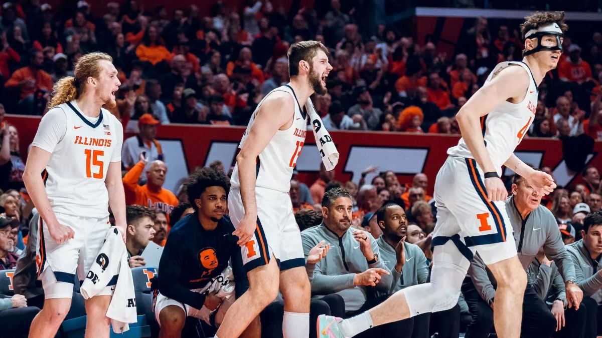 Illinois basketball players in white uniforms cheer on the sideline during a game, crowd in orange in the background.