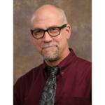 Portrait of a smiling older man with black-framed glasses, wearing a burgundy shirt and paisley tie against a brown studio backdrop.