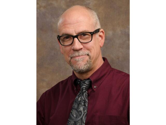 Portrait of a smiling older man with black-framed glasses, wearing a burgundy shirt and paisley tie against a brown studio backdrop.