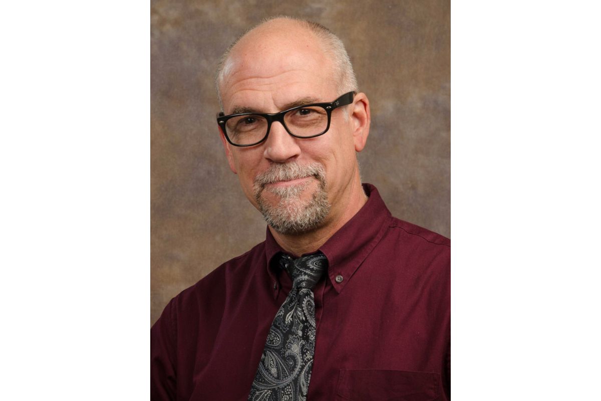 Portrait of a smiling older man with black-framed glasses, wearing a burgundy shirt and paisley tie against a brown studio backdrop.