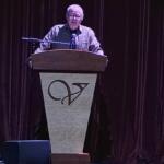 Older man at a wooden podium delivering a speech on a stage with a microphone and dark curtains behind him.