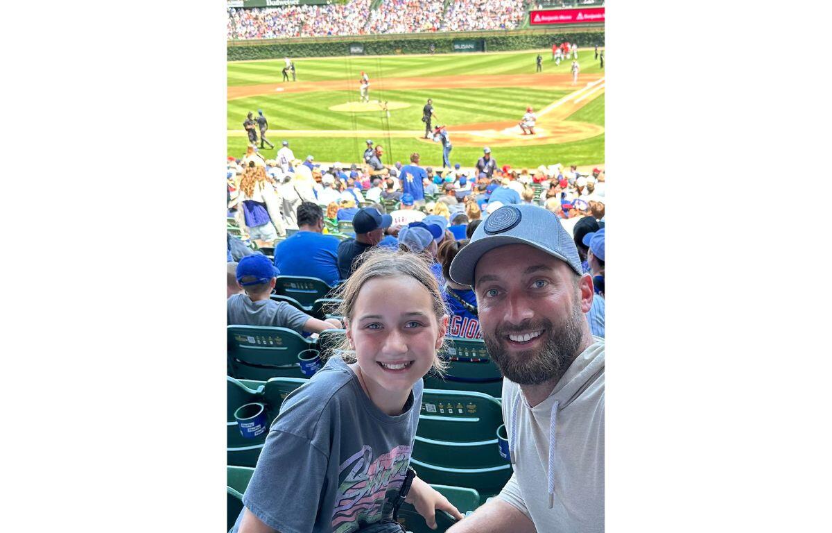 A man and a girl smiling at a baseball game from the stands, with the field and players visible in the background.