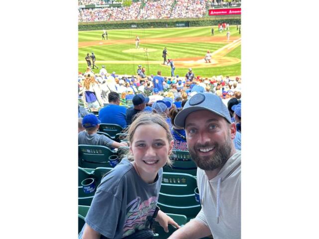 A man and a girl smiling at a baseball game from the stands, with the field and players visible in the background.