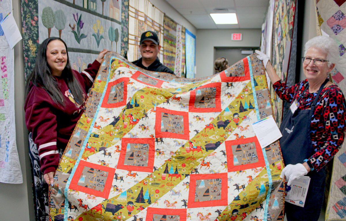 Three people hold a colorful quilt in a hallway, smiling at the camera at a quilting event.