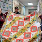 Three people hold a colorful quilt in a hallway, smiling at the camera at a quilting event.