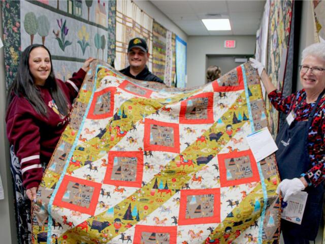 Three people hold a colorful quilt in a hallway, smiling at the camera at a quilting event.