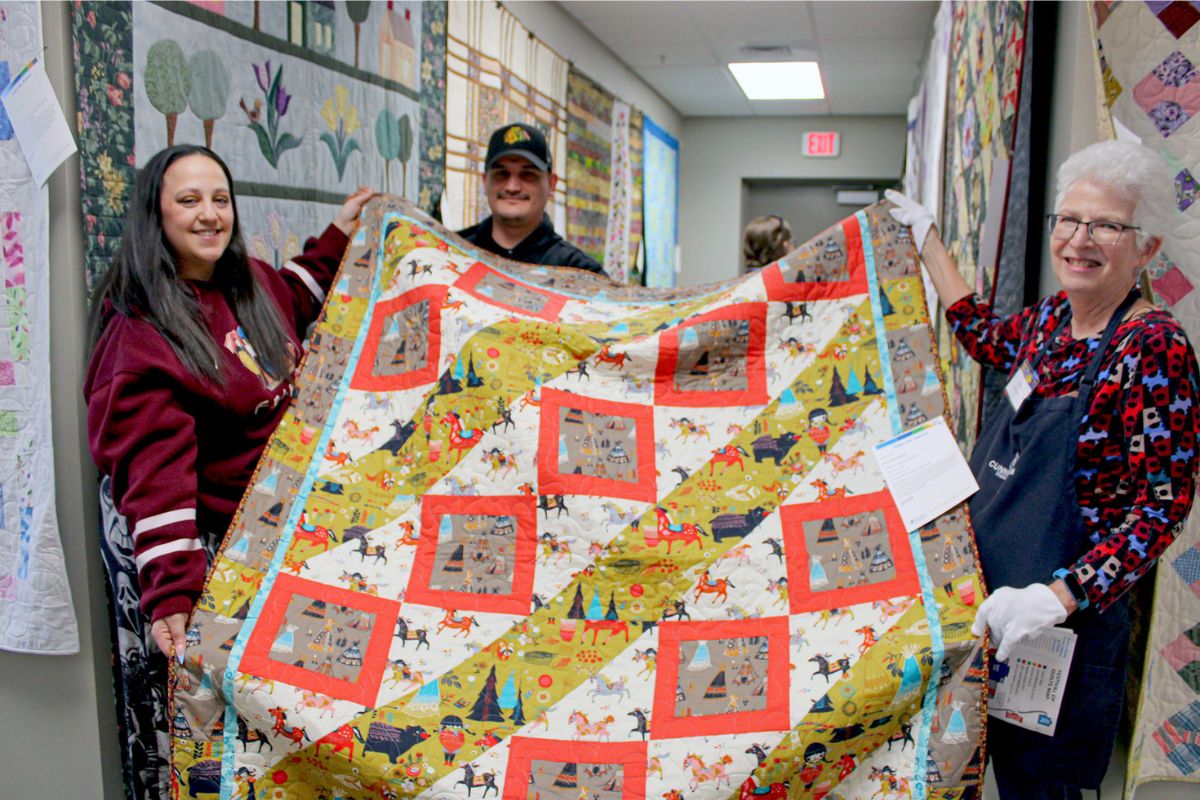 Three people hold a colorful quilt in a hallway, smiling at the camera at a quilting event.