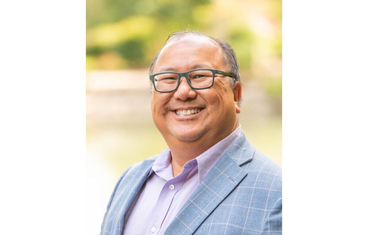 Smiling man with glasses wearing a light blue plaid blazer and lavender shirt outdoors, portrait-style.