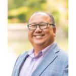 Smiling man with glasses wearing a light blue plaid blazer and lavender shirt outdoors, portrait-style.