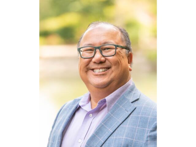 Smiling man with glasses wearing a light blue plaid blazer and lavender shirt outdoors, portrait-style.