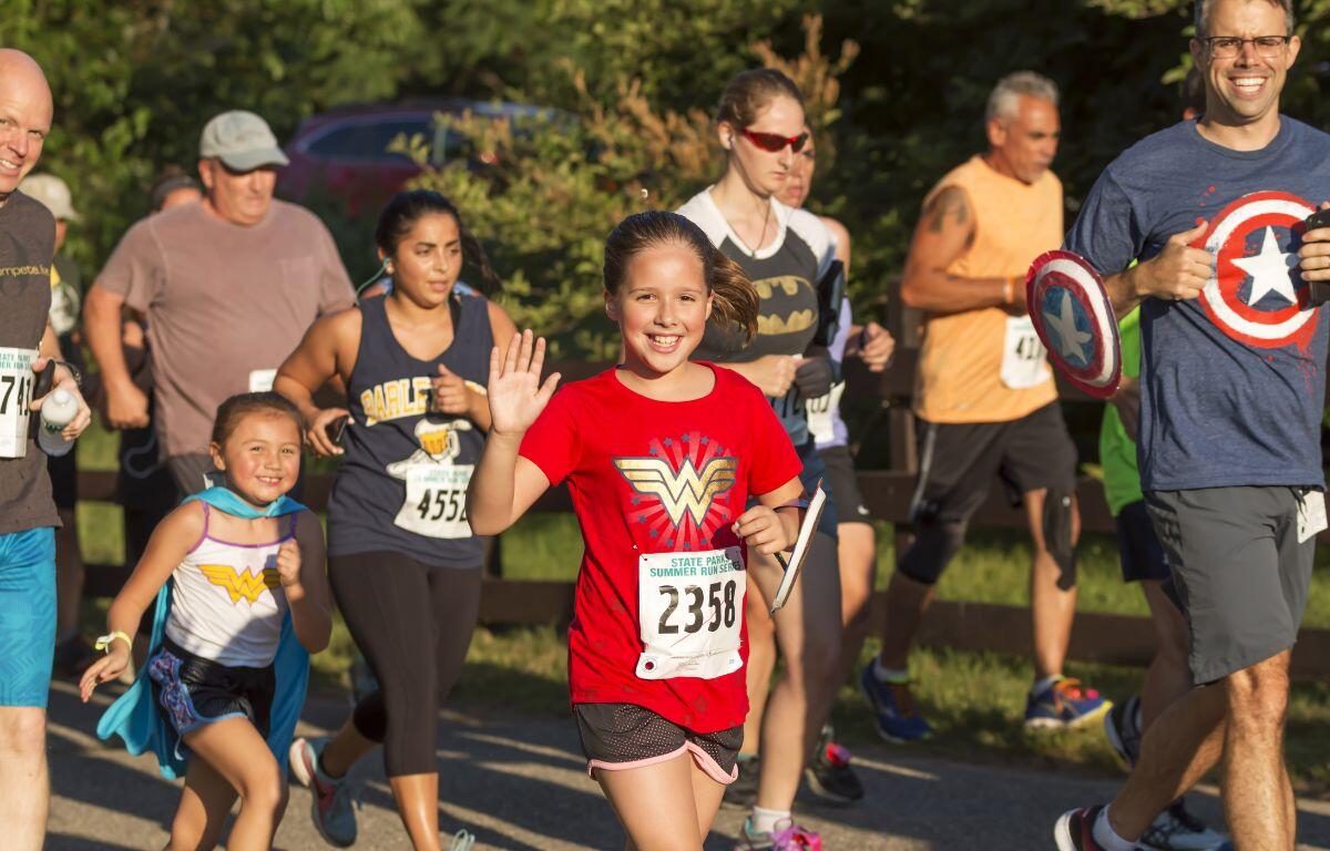 Smiling girl in a red Wonder Woman shirt waves at the camera while running a race; bib 2358 visible on her bib.