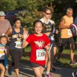 Smiling girl in a red Wonder Woman shirt waves at the camera while running a race; bib 2358 visible on her bib.