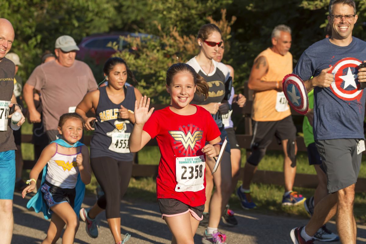 Smiling girl in a red Wonder Woman shirt waves at the camera while running a race; bib 2358 visible on her bib.