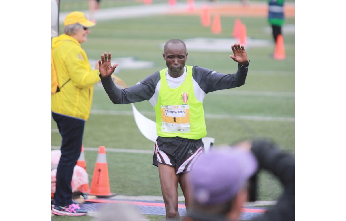 Runner crossing a finish line with arms raised, wearing a neon vest and bib number, on a track field with cones and volunteers nearby.