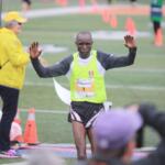 Runner crossing a finish line with arms raised, wearing a neon vest and bib number, on a track field with cones and volunteers nearby.