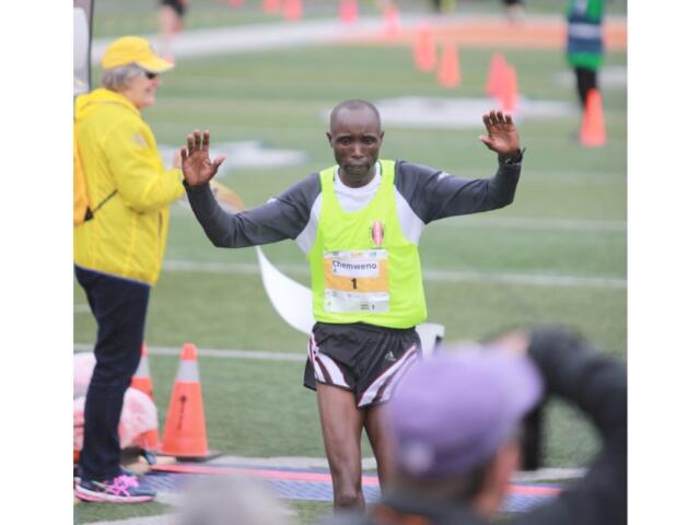 Runner crossing a finish line with arms raised, wearing a neon vest and bib number, on a track field with cones and volunteers nearby.