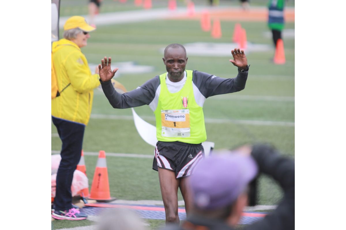 Runner crossing a finish line with arms raised, wearing a neon vest and bib number, on a track field with cones and volunteers nearby.