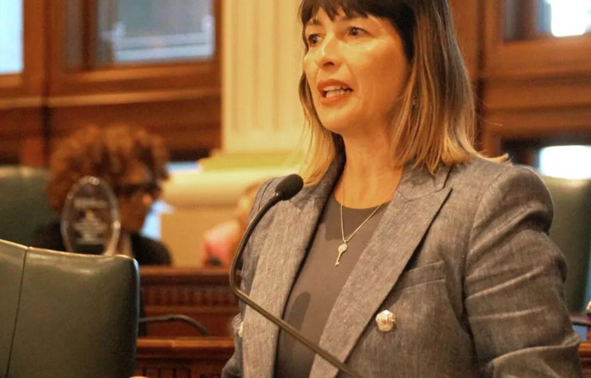 Woman in a gray blazer speaks into a microphone in a formal council chamber.